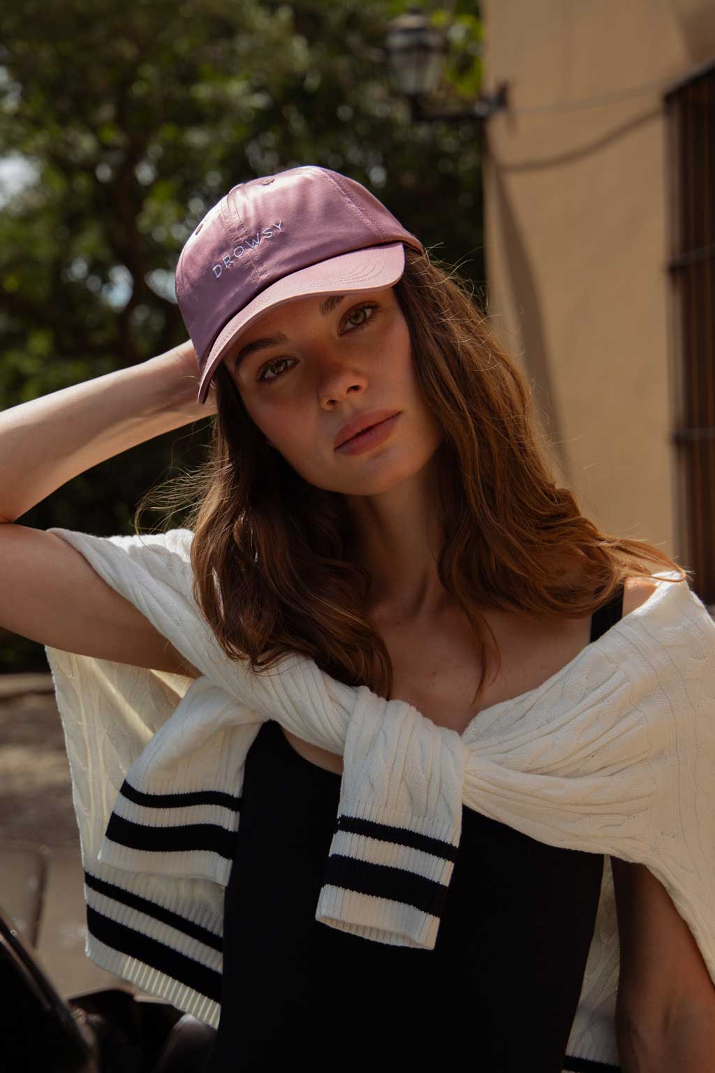 A woman with wavy brown hair wears a mauve baseball cap, a white sweater draped over her shoulders, and a black top. She stands outdoors, hand behind her head, with sunlight and greenery in the background.