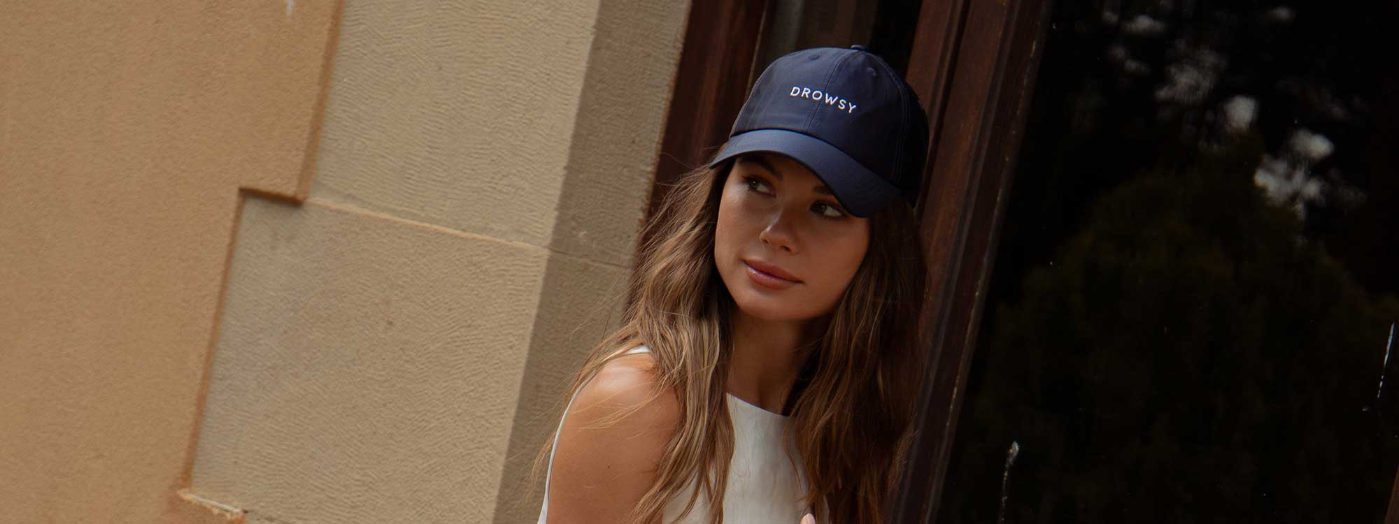A woman with long brown hair, wearing a black baseball cap and a white sleeveless top, stands against a stone wall, looking slightly to her left.