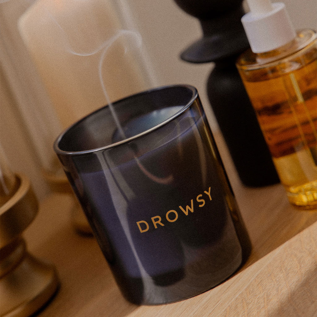 A dark glass Sleep Ritual Candle 220g by drowsysleepco sits on wood, a wisp of smoke curling up—perfect for relaxation or pre-sleep routines. Nearby, a bottle of oil and decorative holders enhance the soothing ambiance.