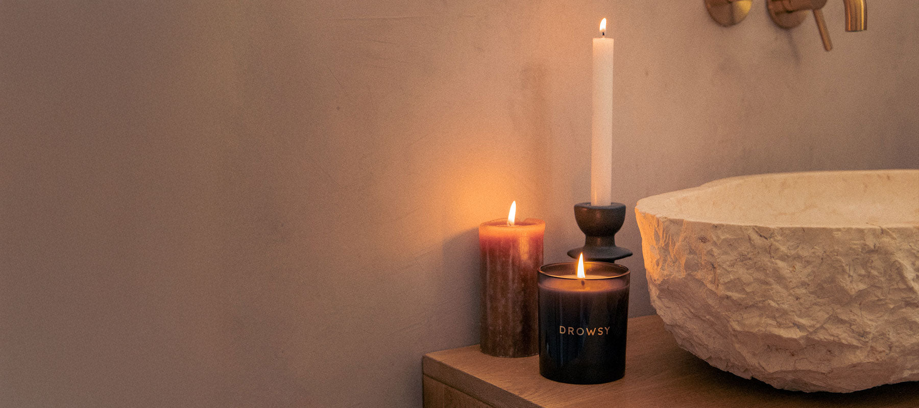 A lit candle labeled DROWSY, a pillar candle, and a white taper candle in a holder sit on a wooden surface next to a textured stone basin against a beige wall.