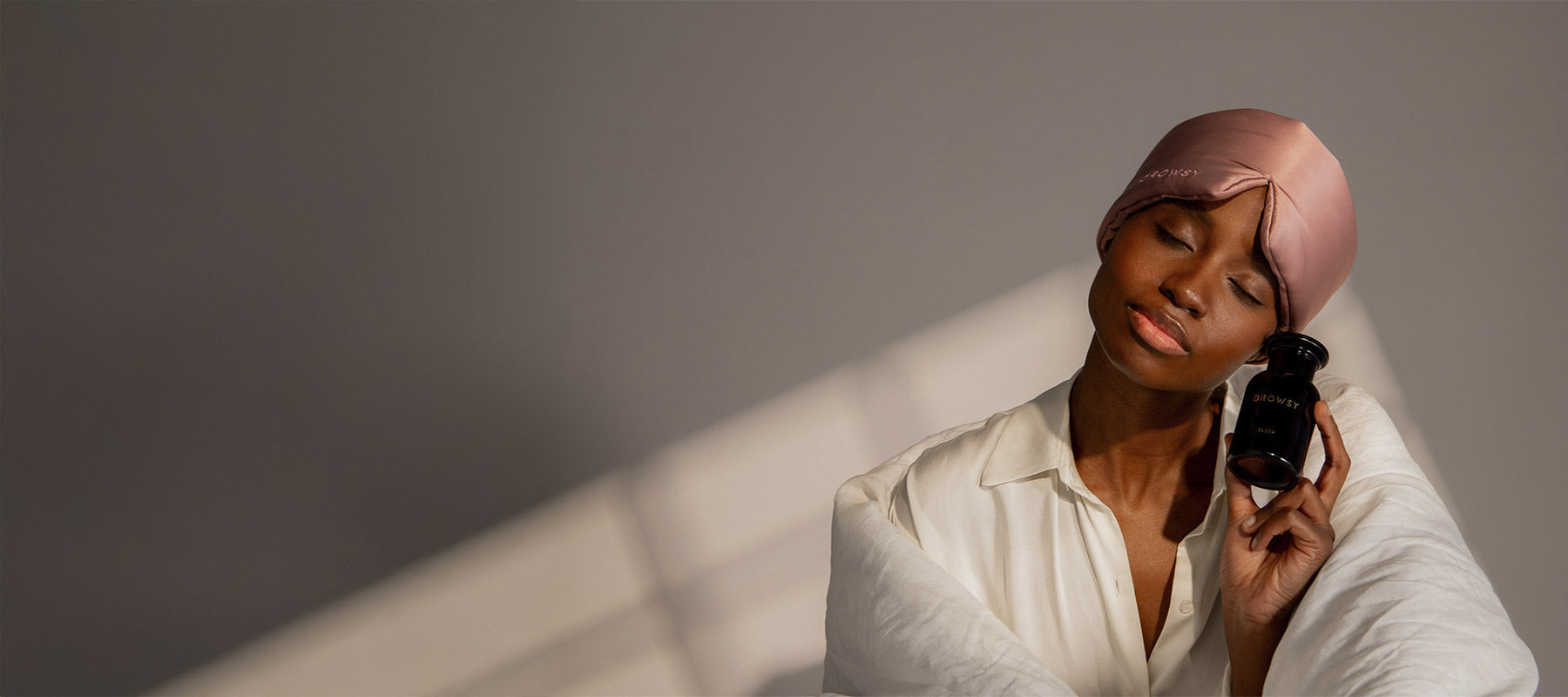 A woman in a silk cap and white shirt closes her eyes and smiles while gently holding a black bottle labeled “pillowtalk” to her cheek, wrapped in a white blanket, with soft light and a neutral background.