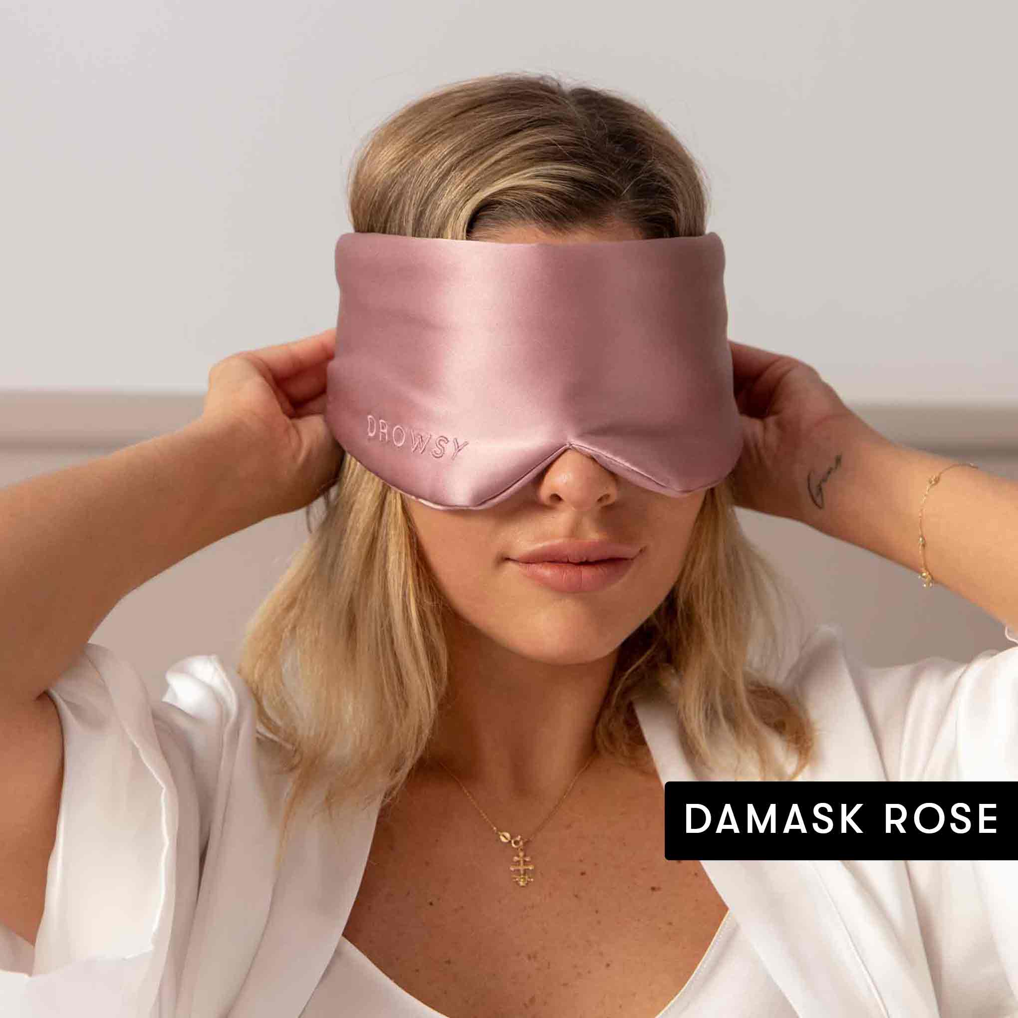 A woman in a white robe with blonde hair and a cross necklace adjusts a Drowsy silk mask from the Lovers Pack over her eyes. DAMASK ROSE appears at the bottom right, showcasing beauty sleep essentials.