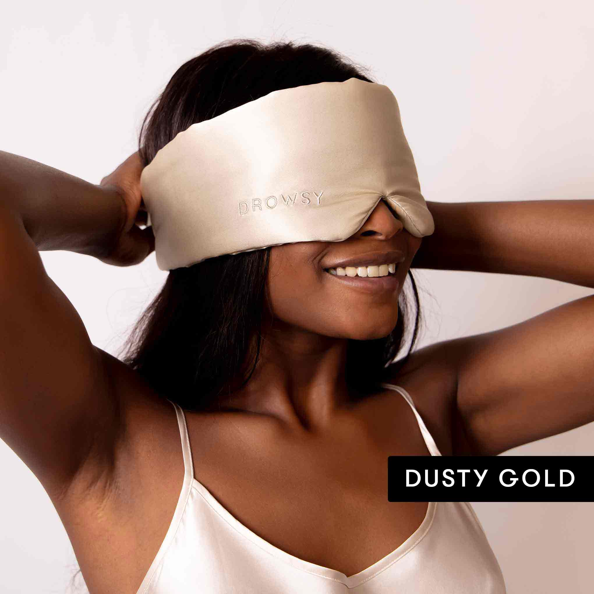 A smiling woman adjusts a Drowsy Besties Pack luxury silk mask over her eyes, wearing a matching silky camisole and ready for beauty sleep. The text on the image reads DUSTY GOLD.