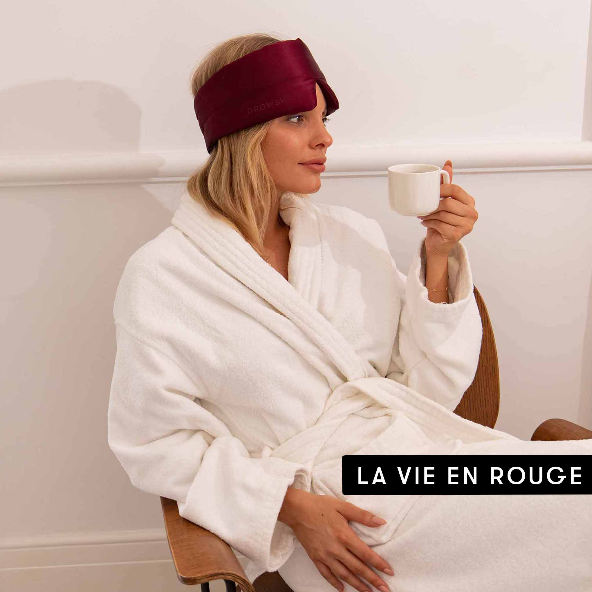A woman in a white bathrobe and maroon Drowsy Besties Pack silk mask sits on a chair with a white mug. The minimal background features LA VIE EN ROUGE text, highlighting beauty sleep in style with Drowsy.