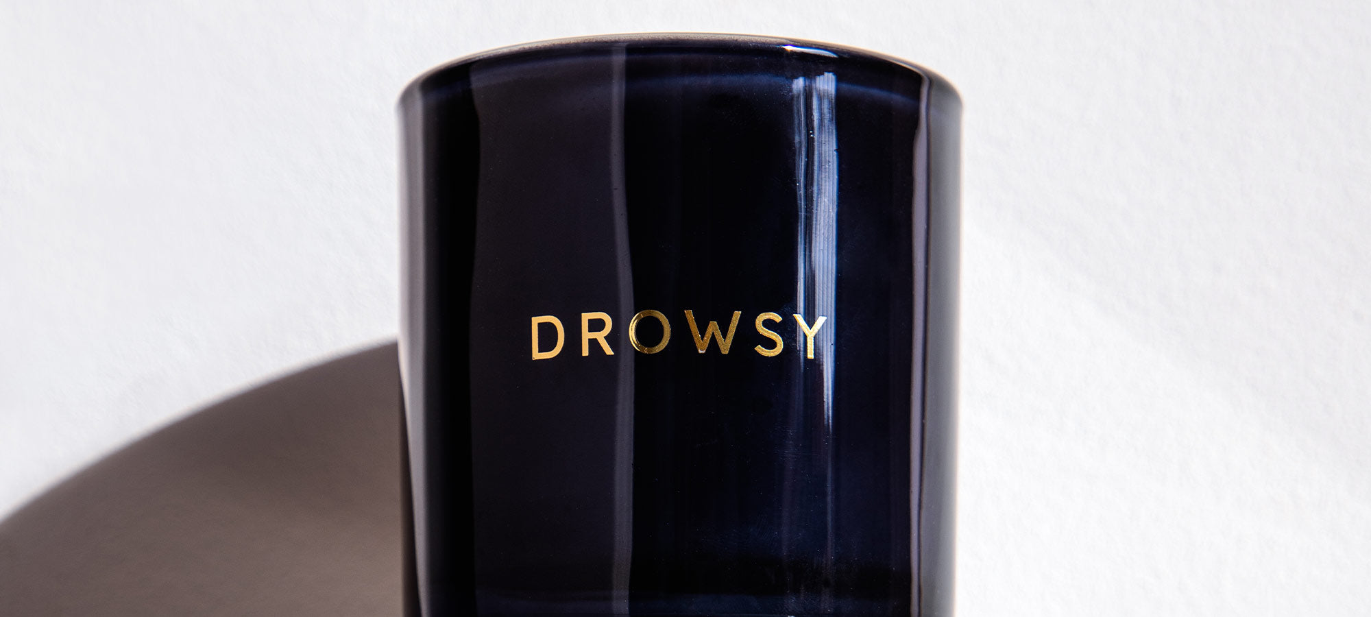 A dark, glossy cup or container with the word DROWSY printed in gold capital letters on the front, placed against a light background with a shadow on the left side.