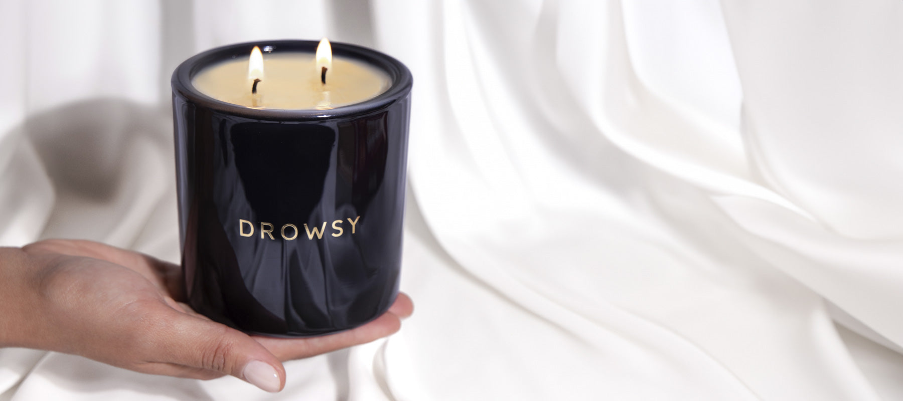A hand holds a black candle jar labeled DROWSY with two burning wicks, against a background of white, softly draped fabric.