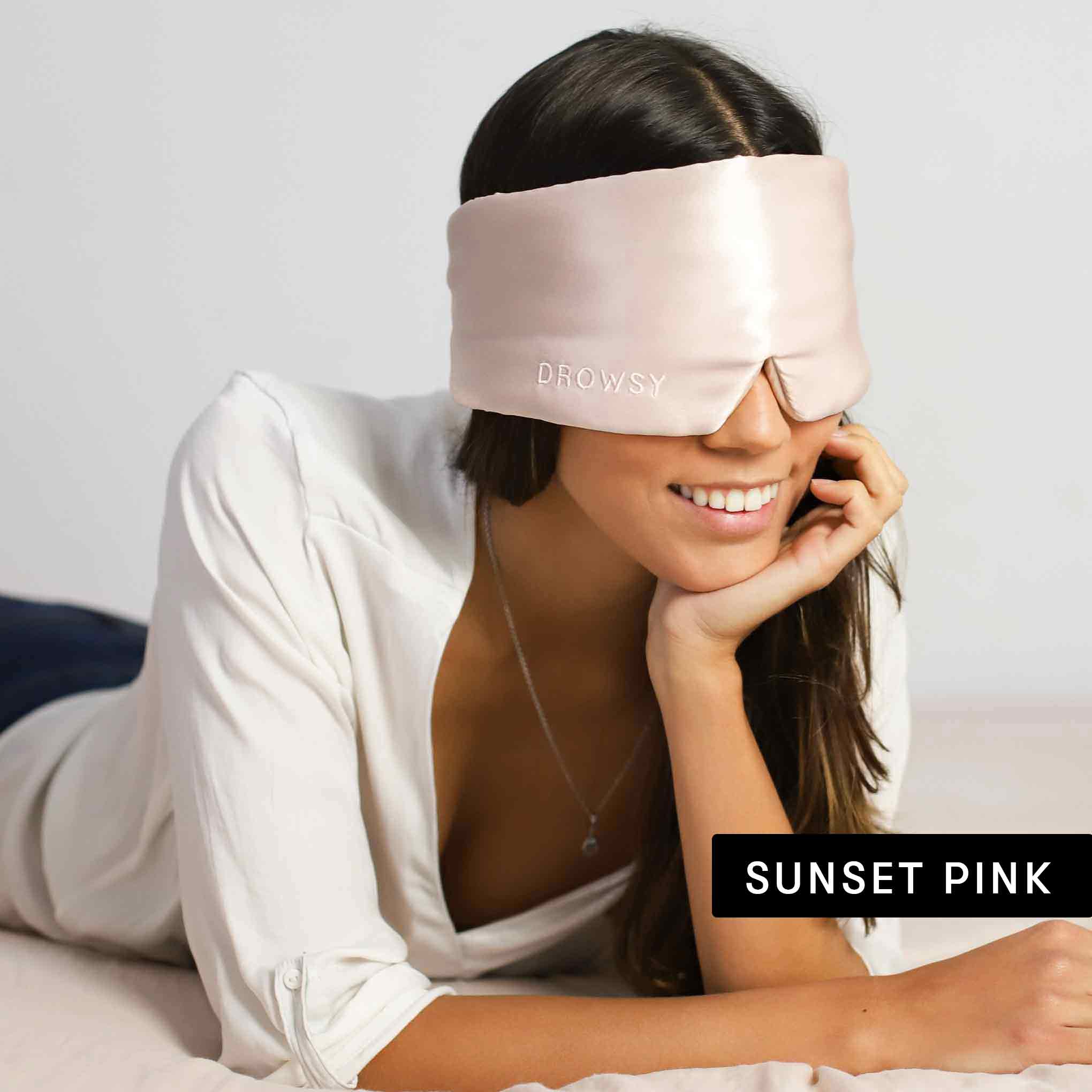 A woman relaxes on a bed wearing a silky light pink Drowsy mask from the Besties Pack. Smiling, she rests her chin on her hand. Sunset Pink appears nearby, promising beauty sleep in mulberry silk.