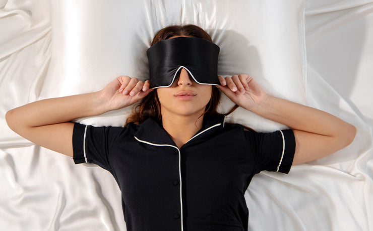 A woman lying on a white silk bed wears a black sleep mask and navy blue pajamas with white trim, holding the mask with both hands while resting on her back.