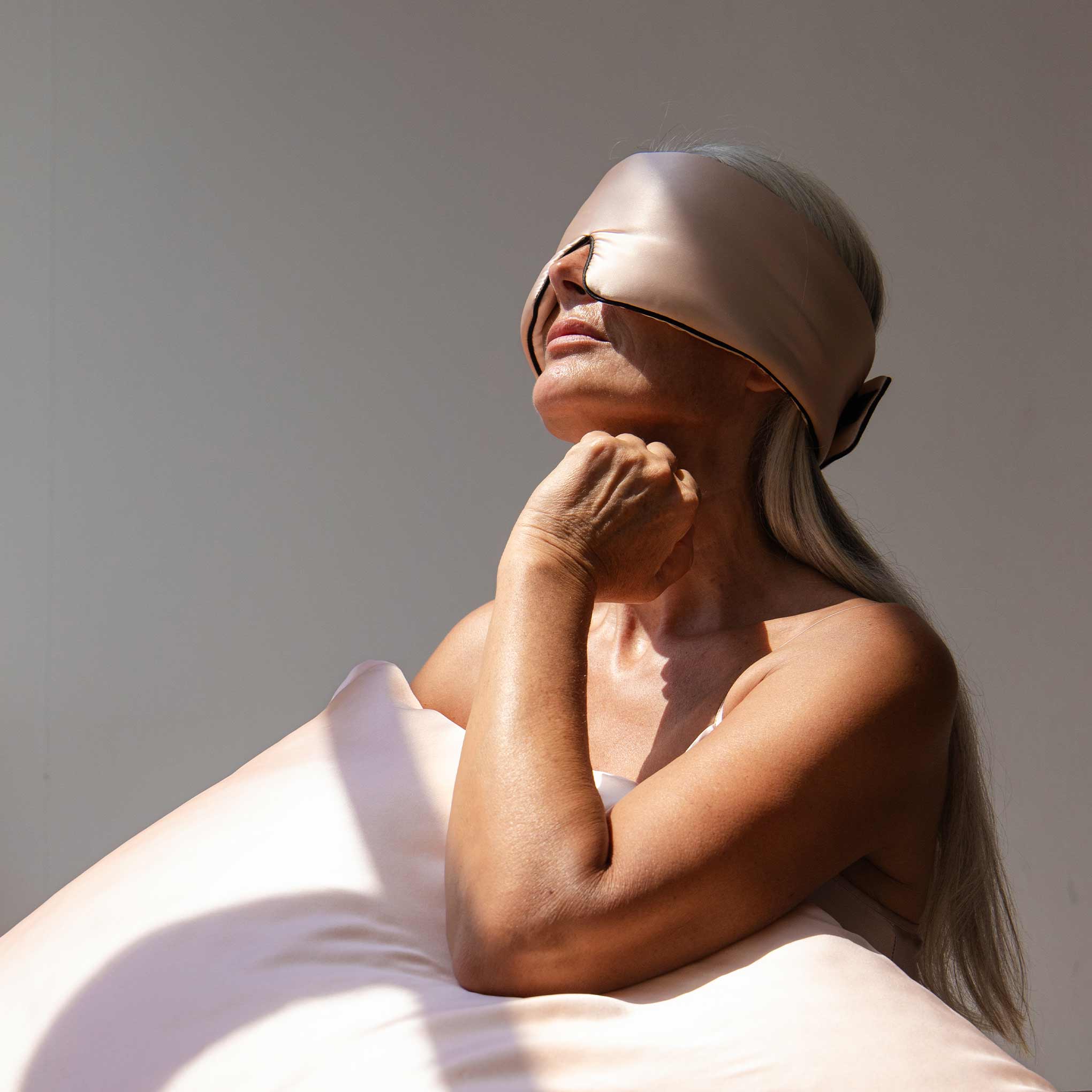A woman with long gray hair wears the us.drowsysleepco Sunset Pink - Pipe Dreams Edition silk sleep mask with adjustable strap, resting her head on a matching satin pillow, bathed in soft light—a perfect moment of beauty sleep.