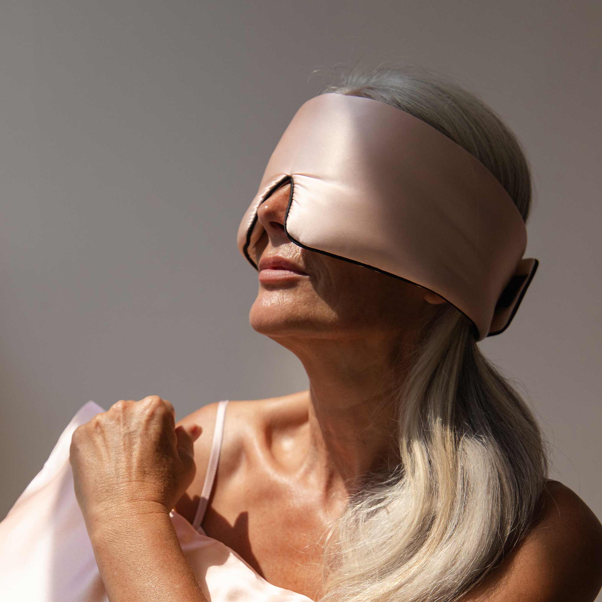 An older woman with long silver hair wears the us.drowsysleepco Sunset Pink - Pipe Dreams Edition silk sleep mask, featuring an adjustable strap and matching pink satin fabric, as she prepares for beauty sleep in soft sunlight.