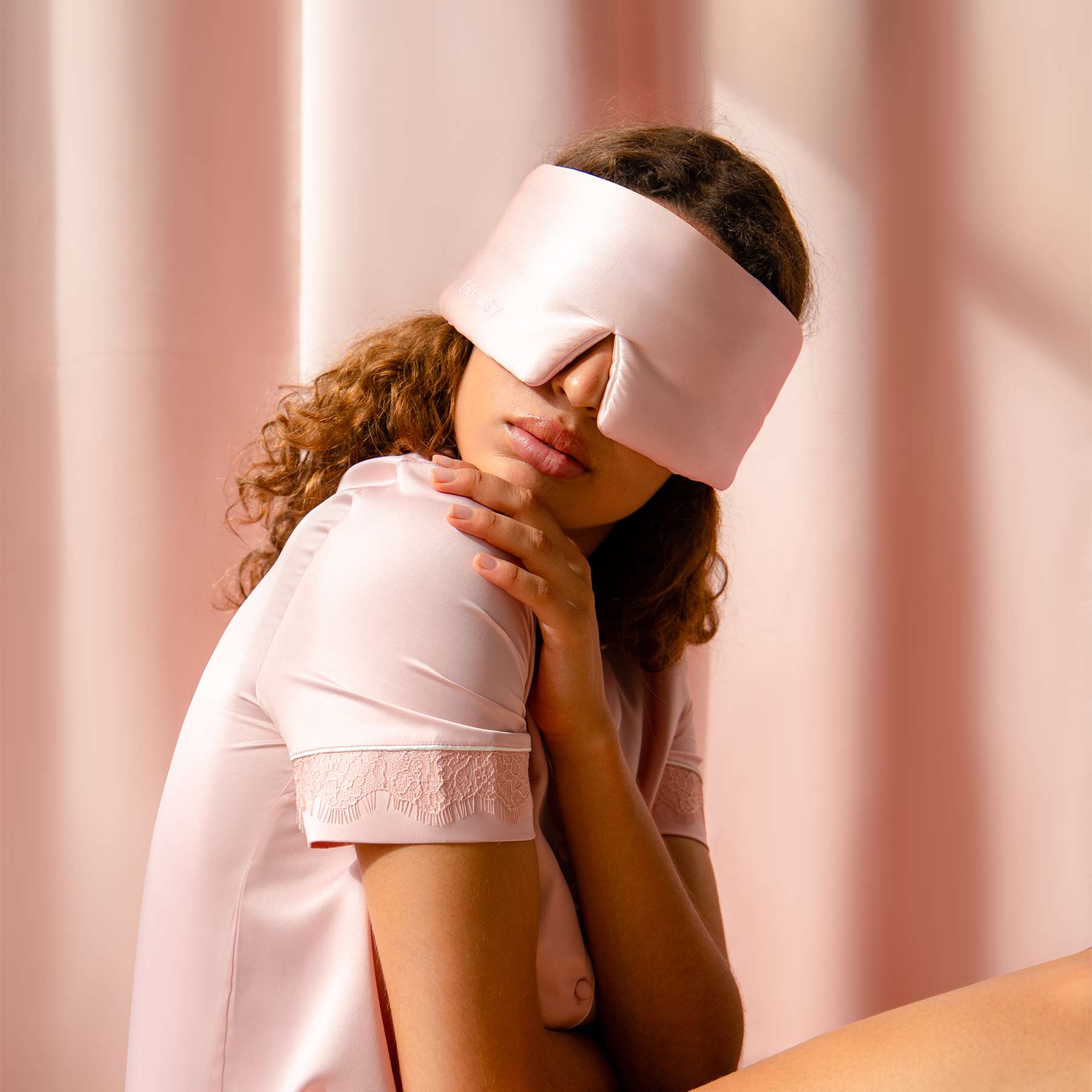 A woman in the Sunset Pink satin sleep mask and matching pajamas by us.drowsysleepco sits with her hand on her shoulder against a soft pink background, softly lit—capturing the essence of beauty sleep.