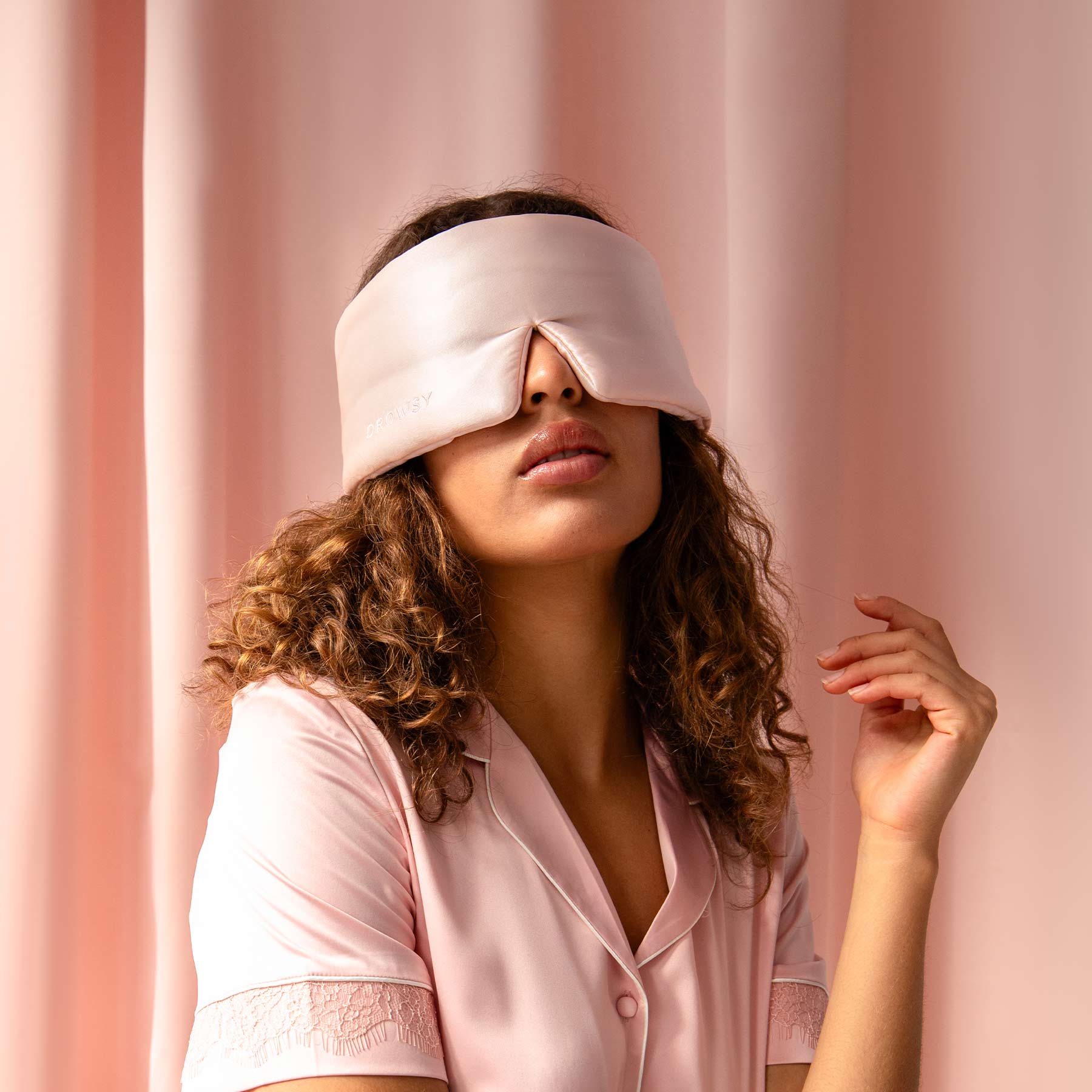 A woman with curly hair in us.drowsysleepco’s Sunset Pink silk eye mask and pajamas sits by matching curtains, preparing for beauty sleep.