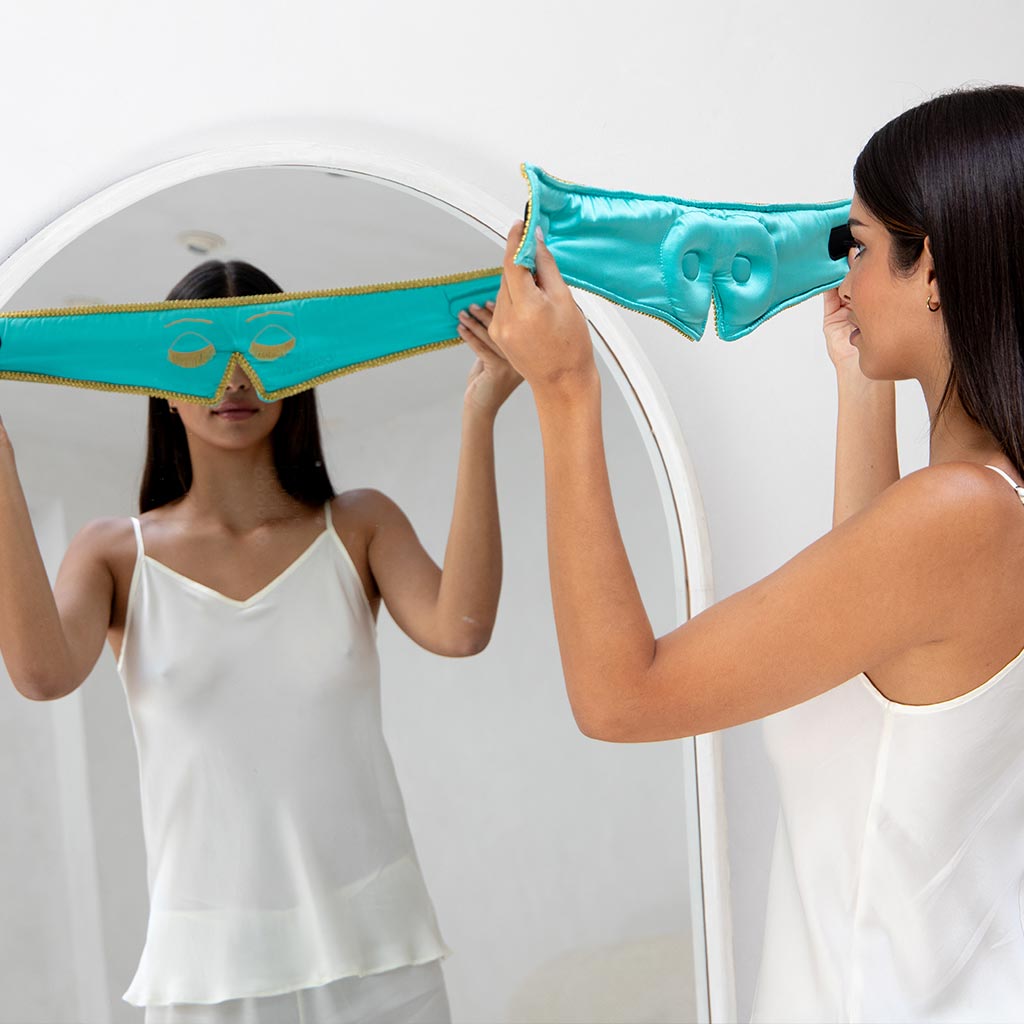A woman in white sleepwear stands before a mirror, holding up the drowsysleepco Breakfast at Drowsy Eyelash Protecting Mask in turquoise satin with gold trim and eye holes, preparing to put it on.