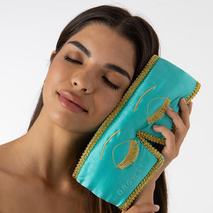 A woman with long dark hair, eyes closed and smiling softly, holds the drowsysleepco Breakfast at Drowsy - Eyelash Protecting Mask—a turquoise and gold embroidered sleep mask—against her cheek. The background is plain white.
