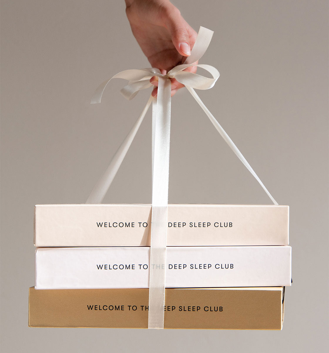 A hand holds three stacked boxes tied with a white ribbon. The boxes are cream, white, and gold, each labeled WELCOME TO THE DEEP SLEEP CLUB on the sides. The background is plain and neutral.