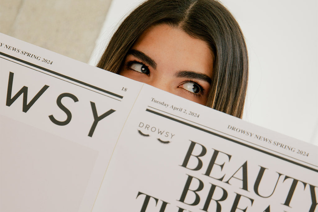 A person with long dark hair looks to the side while holding up two newspapers titled Drowsy News Spring 2024, partially covering their face except for their eyes and forehead.