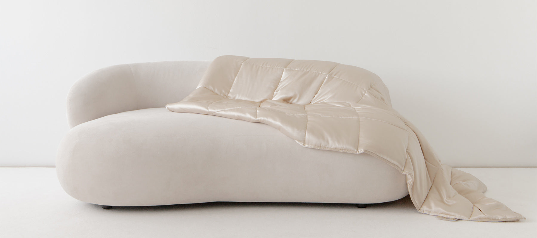 A modern, off-white curved sofa with a shiny beige quilted blanket draped casually over one side, set against a plain white wall and floor.