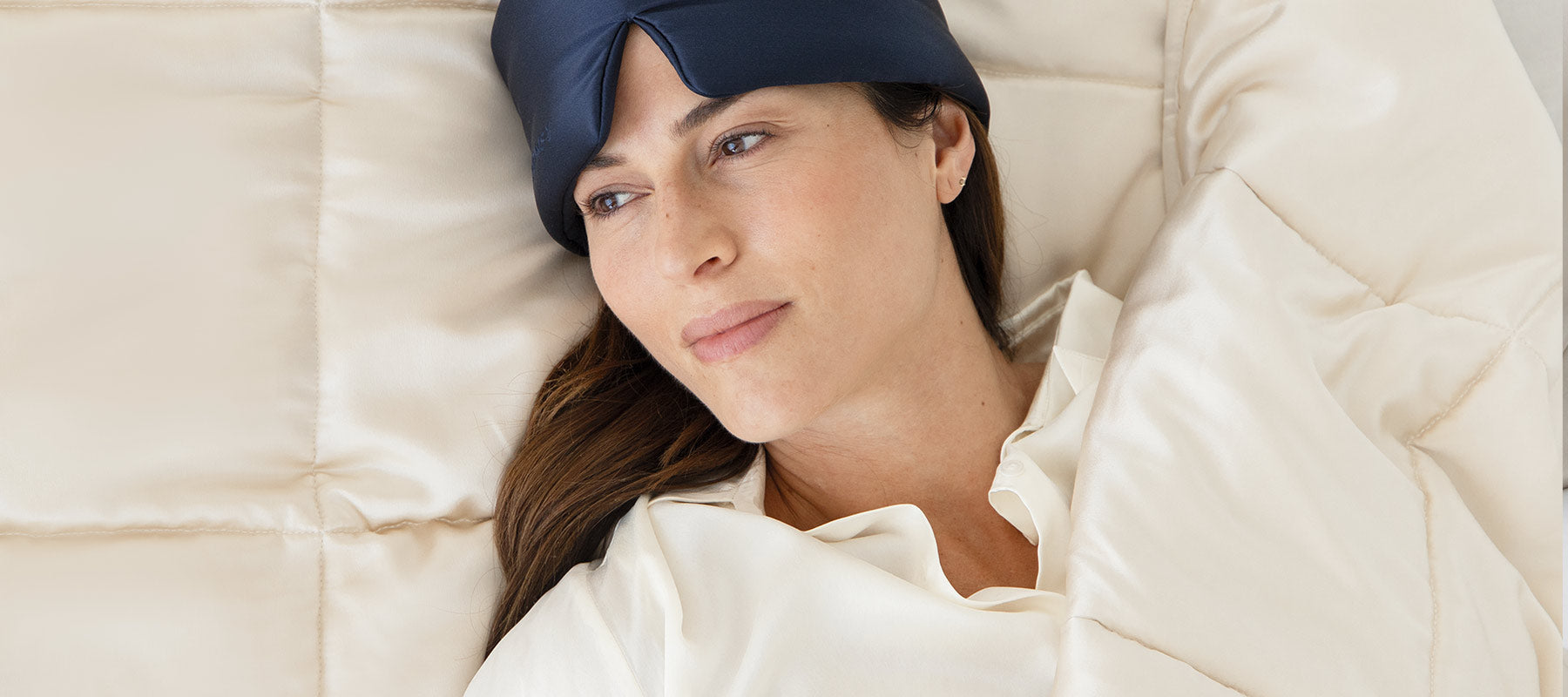 A woman wearing a dark sleep mask pushed up on her forehead lies in bed under a cream-colored comforter, gazing thoughtfully to the side.