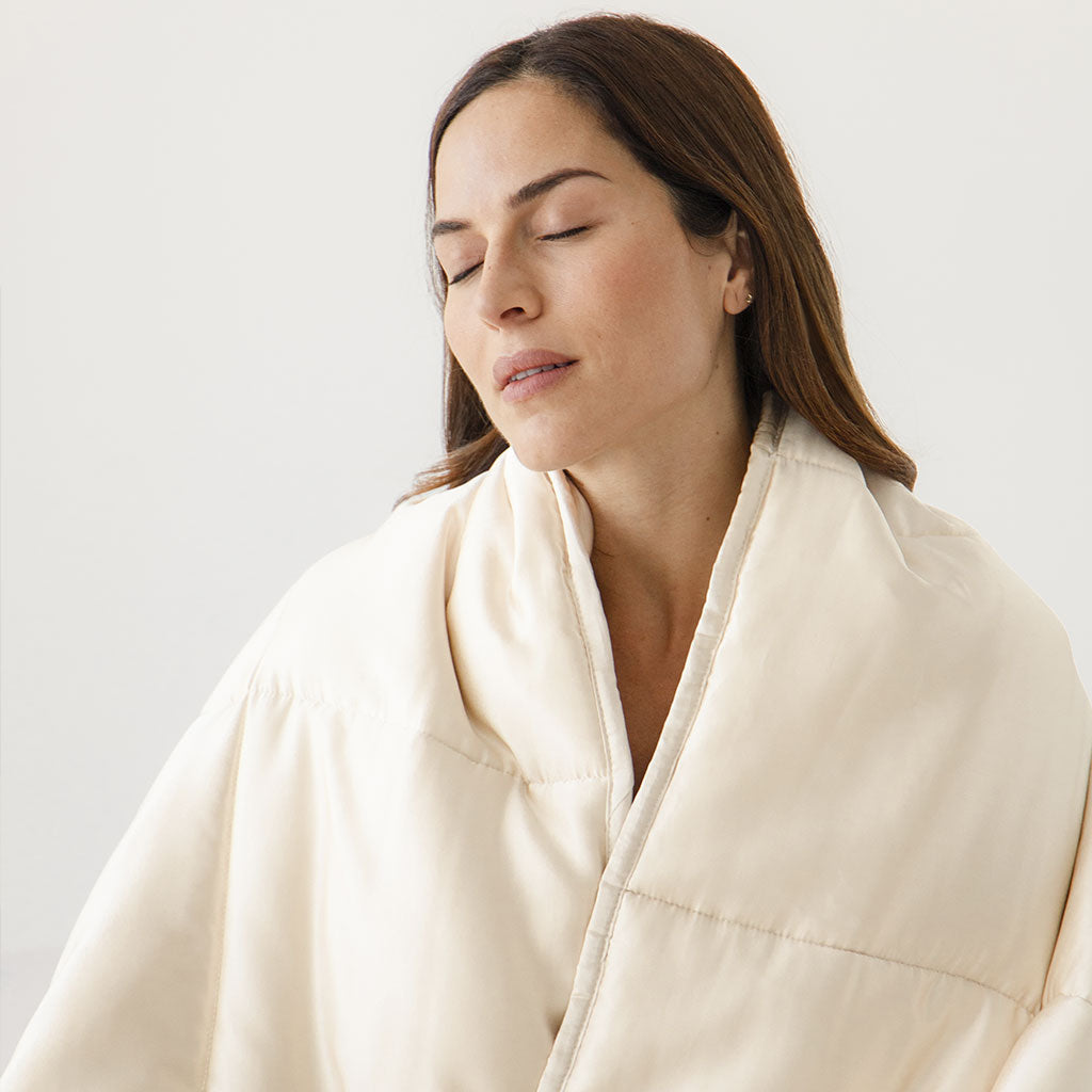 A woman with long brown hair is wrapped in the us.drowsysleepco Dusty Gold Silk Weighted Blanket, eyes closed and peaceful, embodying deep relaxation against a simple light background.