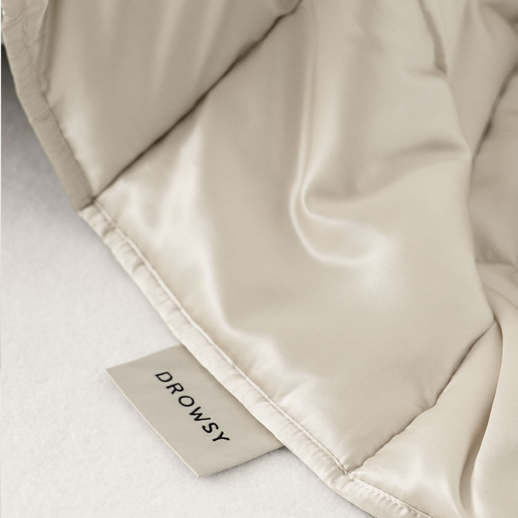 A close-up of the Dusty Gold Silk Weighted Blanket by us.drowsysleepco, featuring a visible DROWSY fabric tag on a soft white surface, offers deep relaxation and an unmatched sleeping experience.