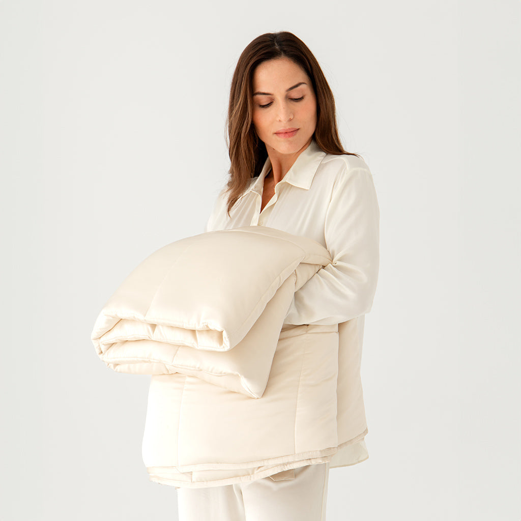 A woman in cream pajamas gently holds the us.drowsysleepco Dusty Gold Silk Weighted Blanket, its light beige silk cover folded neatly, against a plain white background. She looks down at it, exuding calm and deep relaxation.