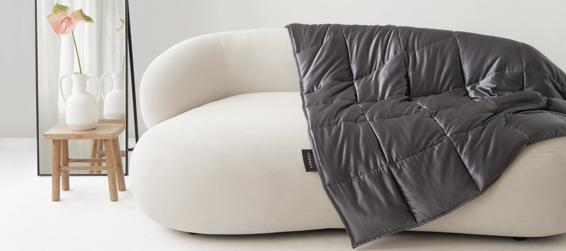 A modern white sofa with a dark quilted blanket draped over one side. Next to the sofa is a small wooden stool holding two white vases, in front of a large floor mirror with a pink flower. The setting is minimal and bright.