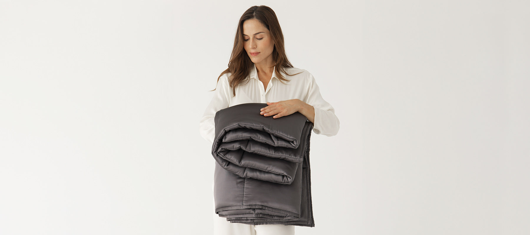 A woman in white pajamas stands against a plain background, holding a neatly folded dark gray comforter in her arms while looking down at it with a gentle expression.