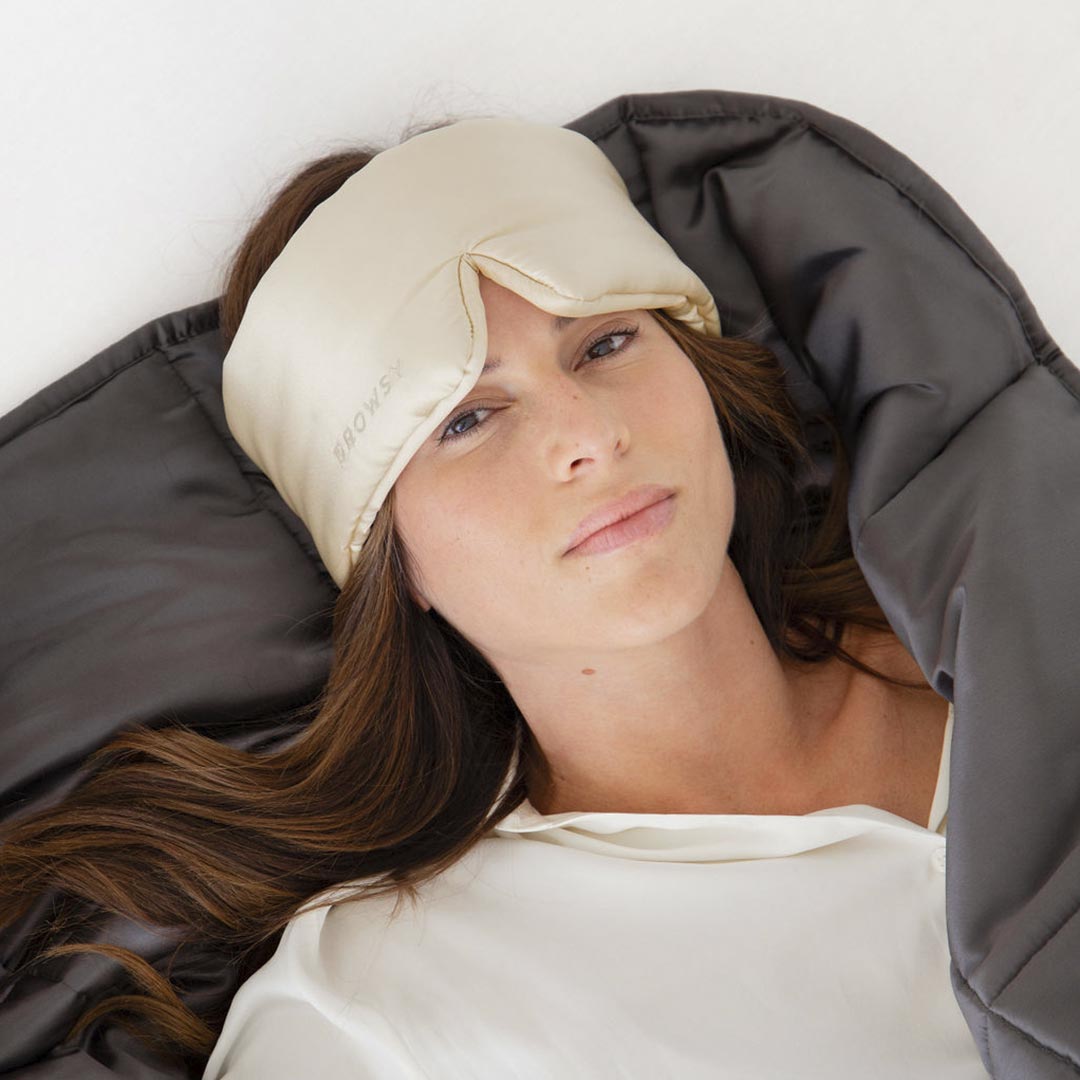 A woman with brown hair relaxes on a bed under the us.drowsysleepco Moonlight Shadow Silk Weighted Blanket in dark grey, wearing a cream sleep mask and white top, looking peaceful and deeply relaxed.