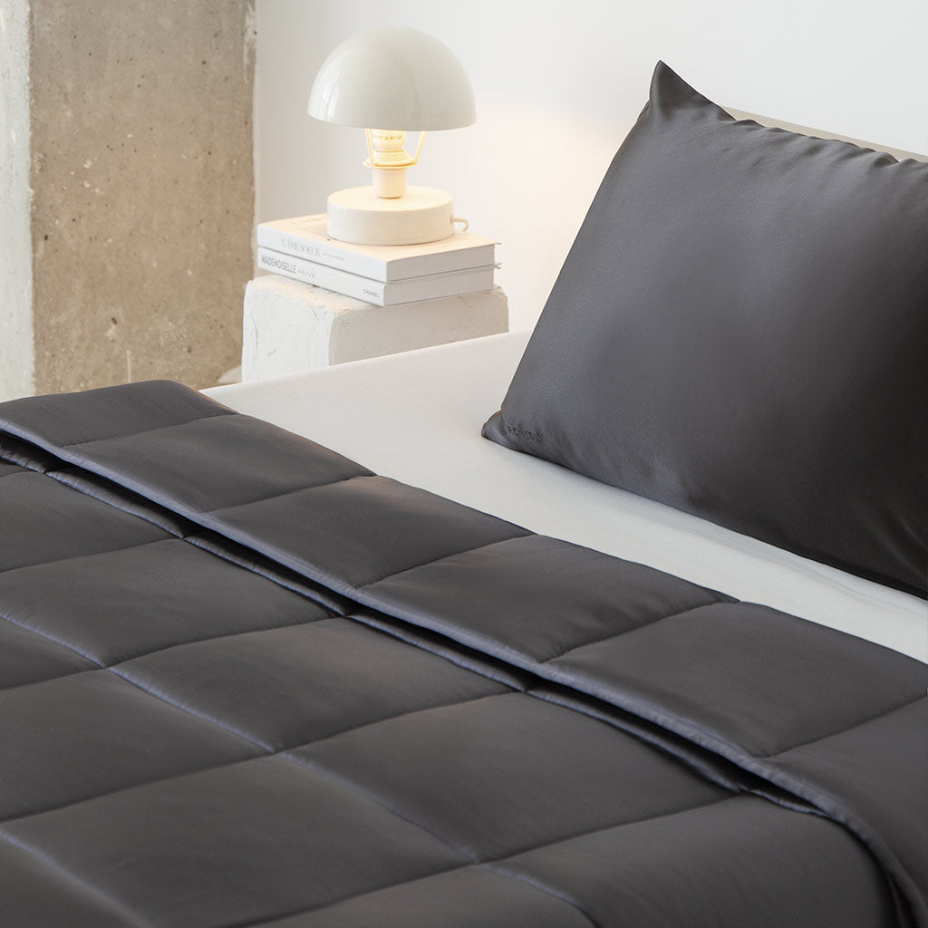 A neatly made bed features the us.drowsysleepco Moonlight Shadow Silk Weighted Blanket in dark gray, paired with a matching pillow. A white lamp rests on stacked books atop a minimalist nightstand beside a concrete wall.