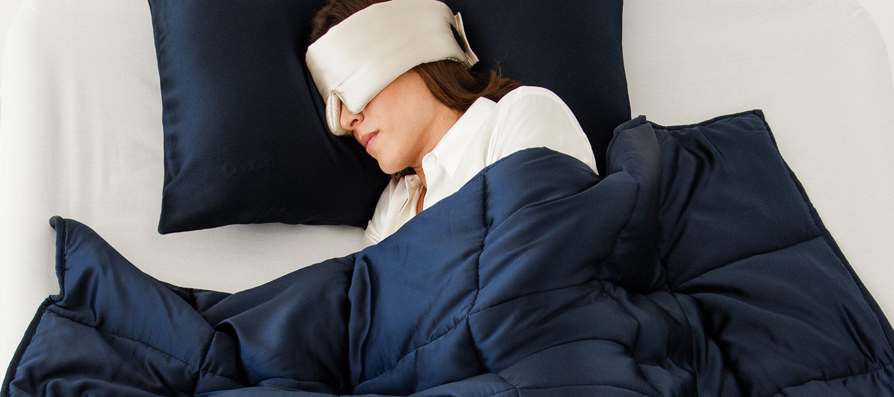 A person wearing a white sleep mask and white shirt is lying in bed under a dark blue comforter, resting their head on a matching blue pillow. The person appears to be asleep.