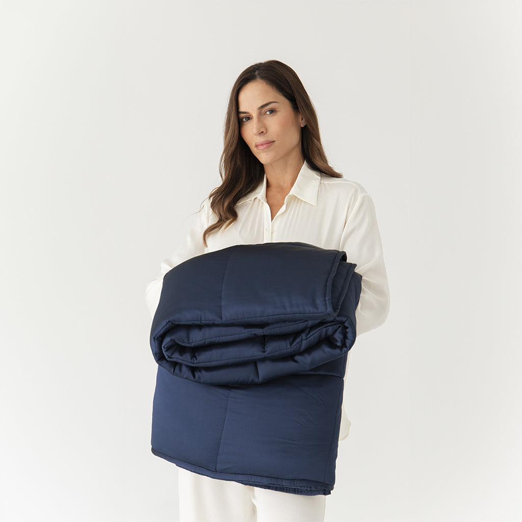 A woman in white holds the us.drowsysleepco Midnight Blue Silk Weighted Blanket, neatly folded, against a plain white background—evoking deep relaxation.