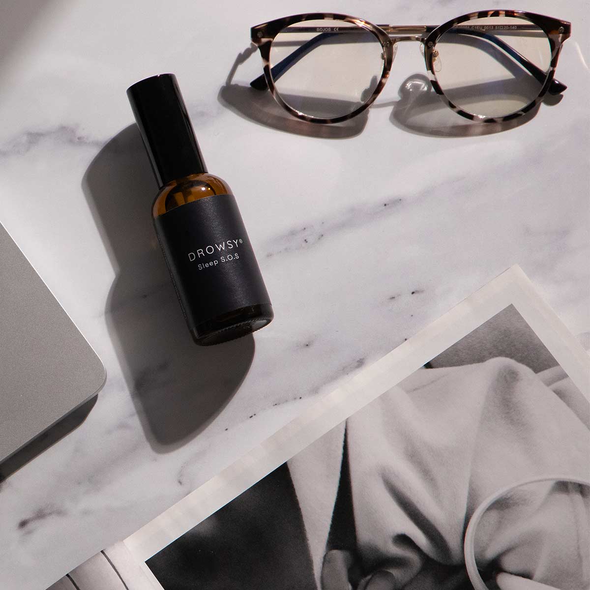 A bottle labeled Drowsy Sleep SOS lies on a marble surface next to tortoiseshell glasses, part of a laptop, and a black-and-white magazine or photo.