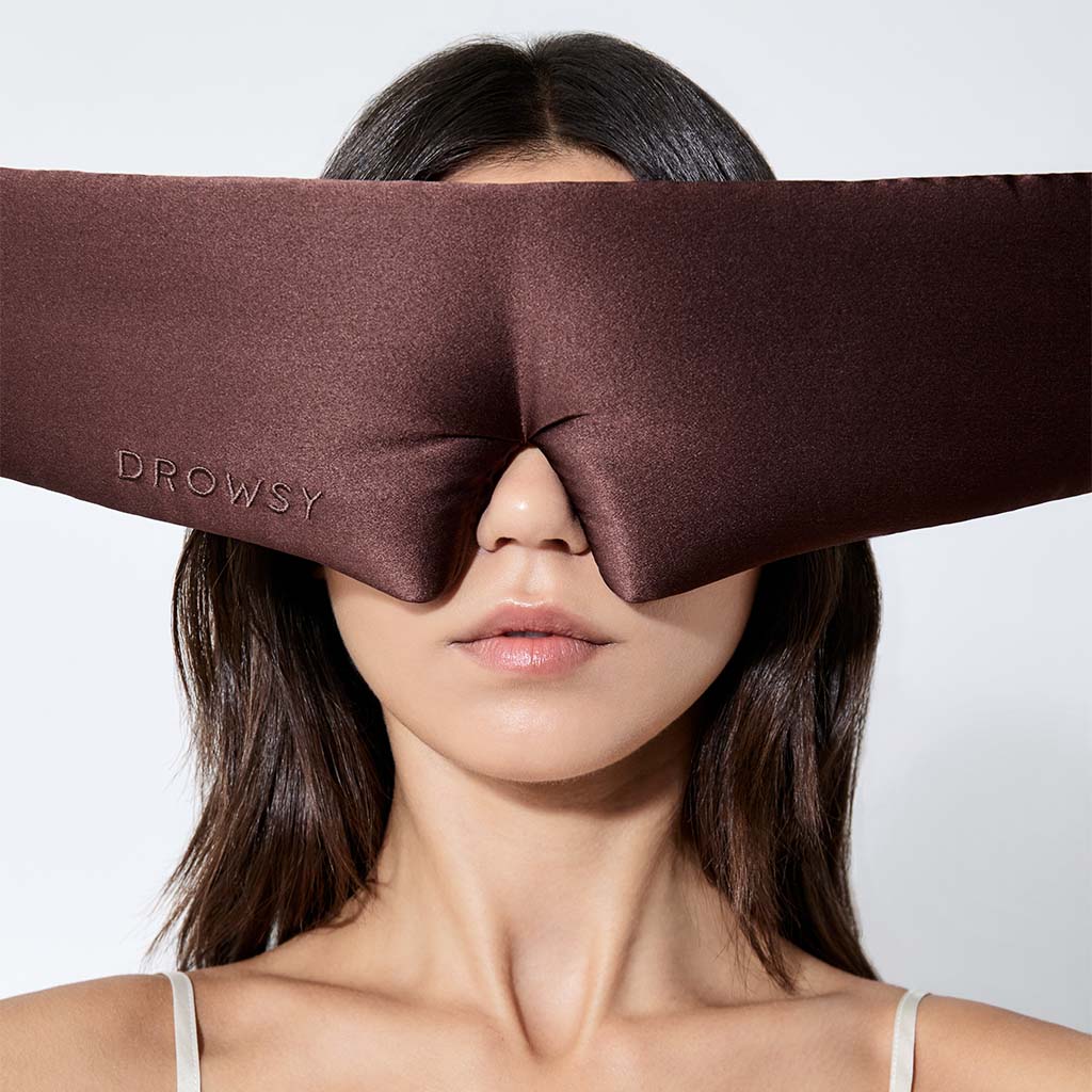 A woman with straight brown hair wears a large brown silk sleep mask labeled 