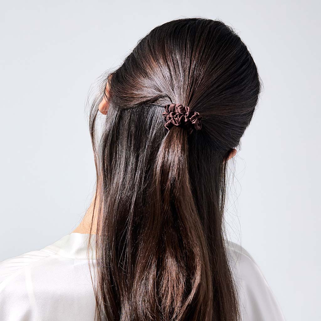 A woman with long, straight brown hair, seen from behind in a white top, styles her hair using the drowsysleepco Skinny Scrunchie Set - Cocoa Glow for gentle and protective hold.