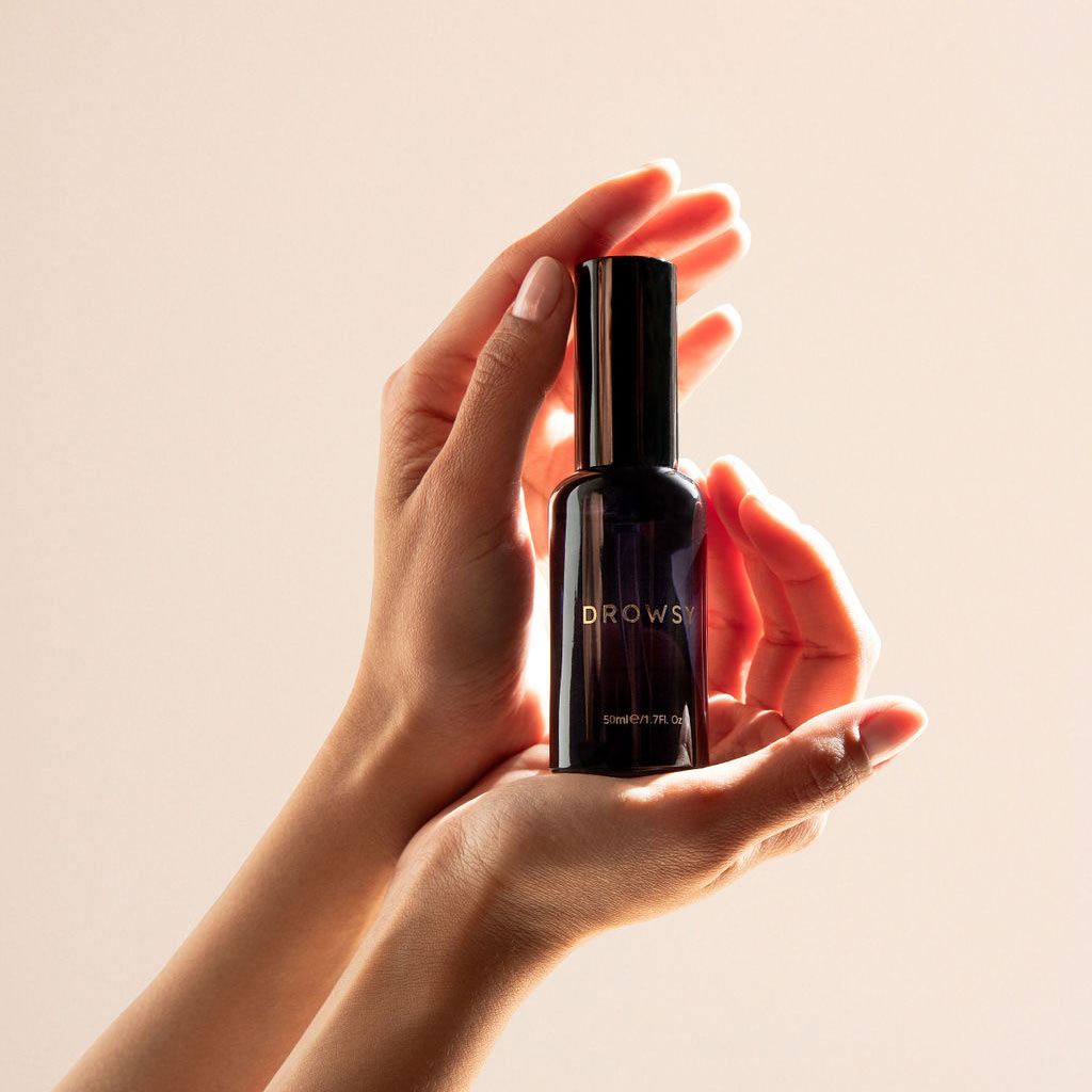 Two hands gently hold a dark glass bottle labeled drowsysleepco Sleep S.O.S Pillow Spray—an inviting addition to your nightly routine—against a soft, neutral background, with light highlighting the bottle and hands.