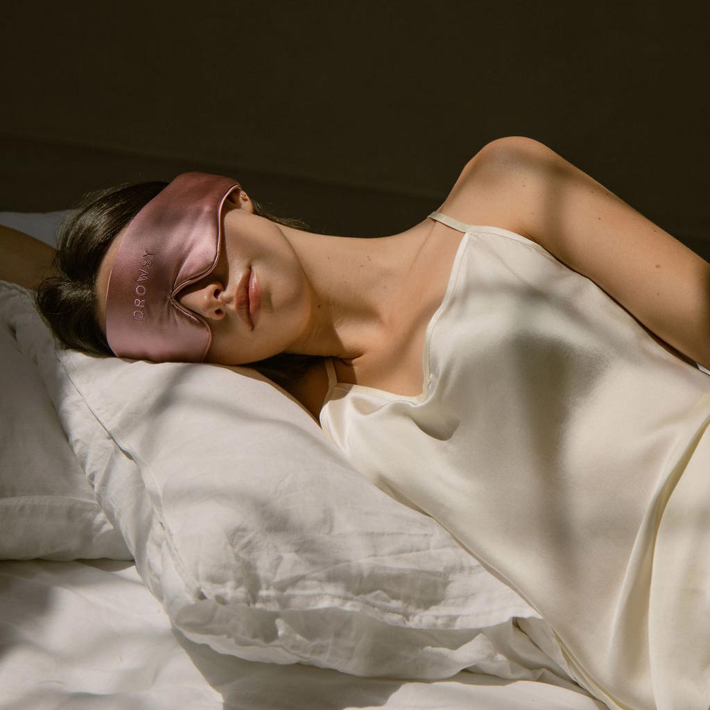 A woman in a white satin camisole lies on white bedding, wearing a pink silk sleep mask. She is resting with one arm above her head, bathed in soft natural light.