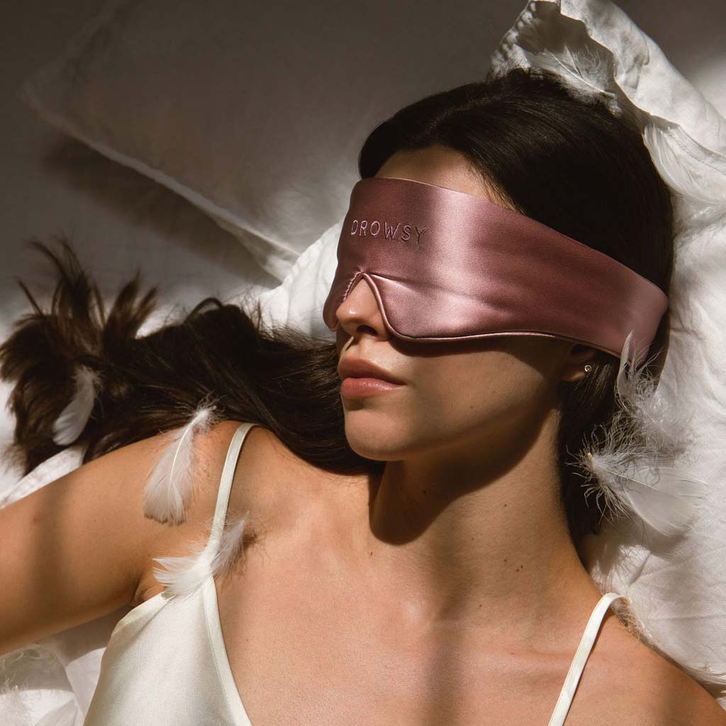A woman lies on a bed wearing a silky pink sleep mask with the word DROWSY on it. She is dressed in a white camisole, surrounded by white pillows and feathers.