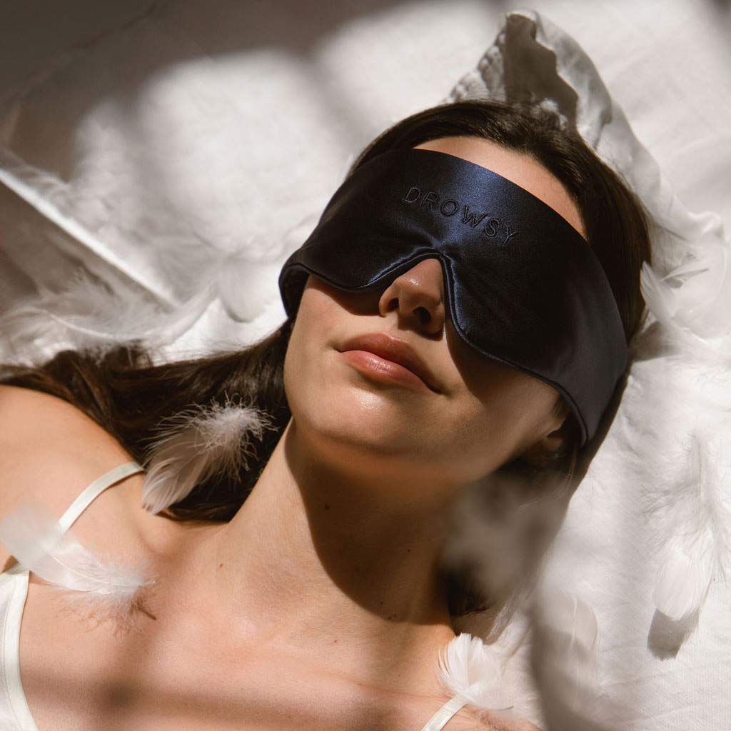 A woman lies on white sheets wearing a black silk sleep mask that says DROWSY. Soft sunlight and scattered white feathers surround her, creating a calm, restful atmosphere.