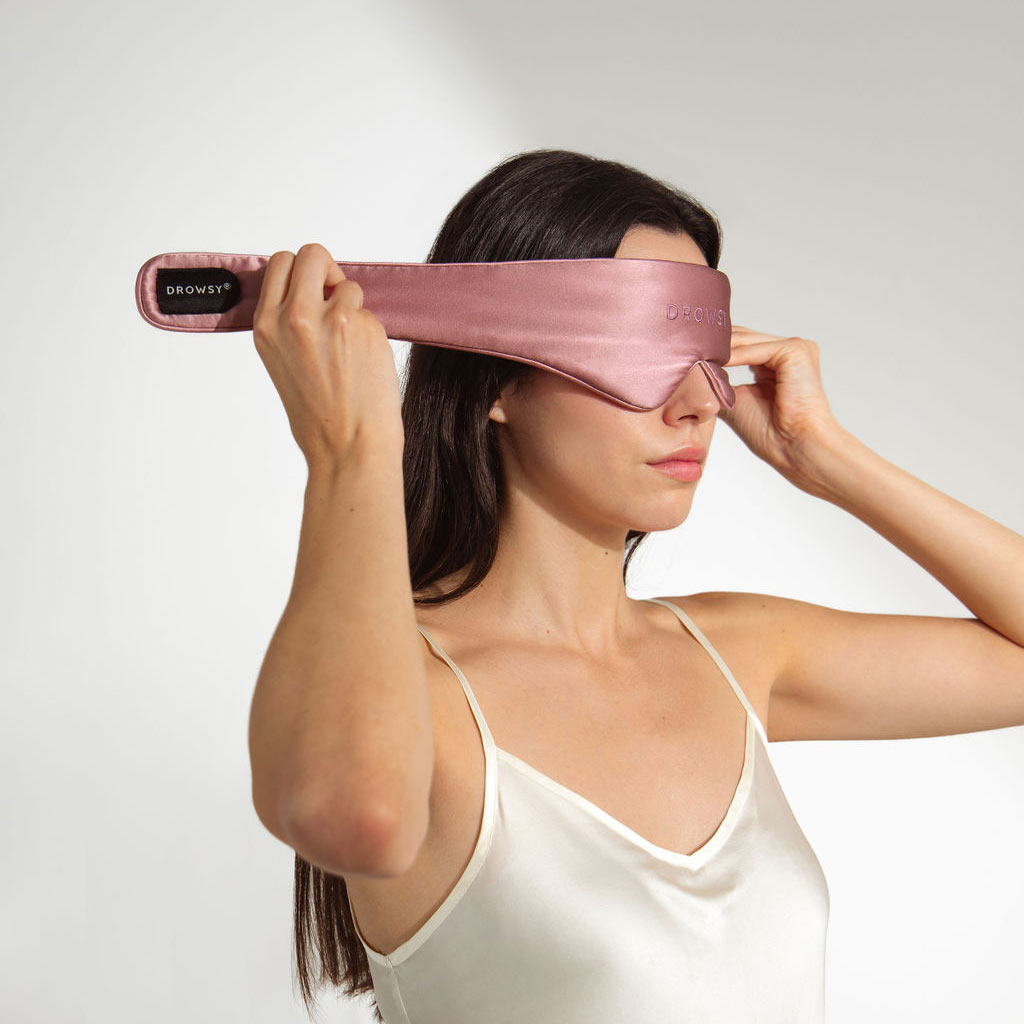 A woman in a white satin camisole puts on the us.drowsysleepco Featherweight Silk Eye Mask Damask Rose, which features a total black-out design, while standing against a plain white background.
