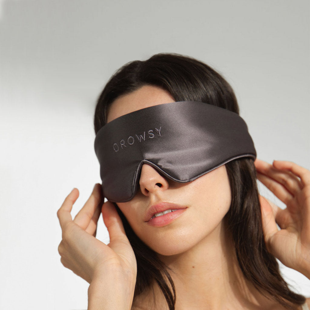 A woman adjusts a silky dark gray sleep mask labeled DROWSY over her eyes against a plain light background.