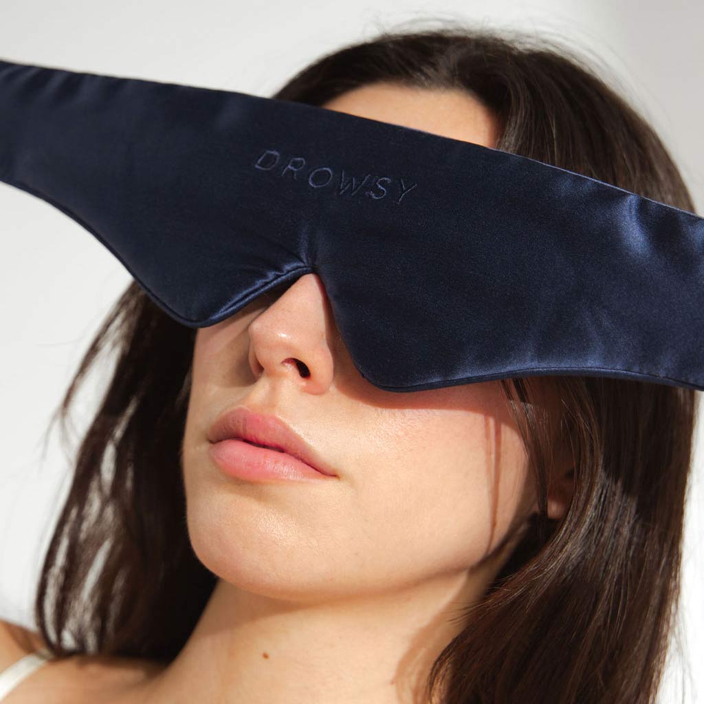 A woman with long brown hair wears a large, dark blue silk sleep mask labeled “DROWSY” that covers her eyes and part of her face. The background is plain and well-lit.