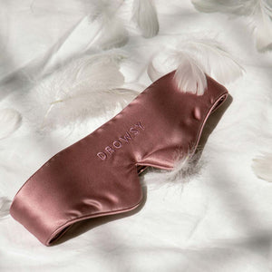 The Featherweight Silk Eye Mask Damask Rose by us.drowsysleepco, featuring a total black-out design and DROWSY embroidery, rests on white fabric and is surrounded by soft white feathers.