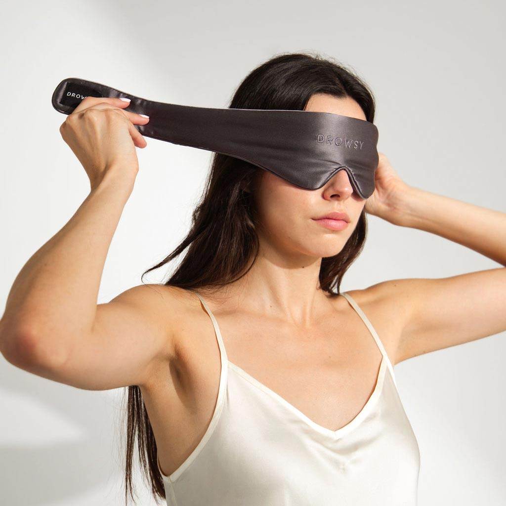 A woman in a cream camisole wears the us.drowsysleepco Featherweight Silk Eye Mask Moonlight Shadow, which offers total blackout, against a plain white background.