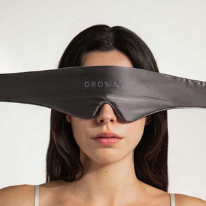A woman with straight dark hair and a neutral expression wears the us.drowsysleepco Featherweight Silk Eye Mask Moonlight Shadow, a wide silky black mask covering her eyes and upper nose.