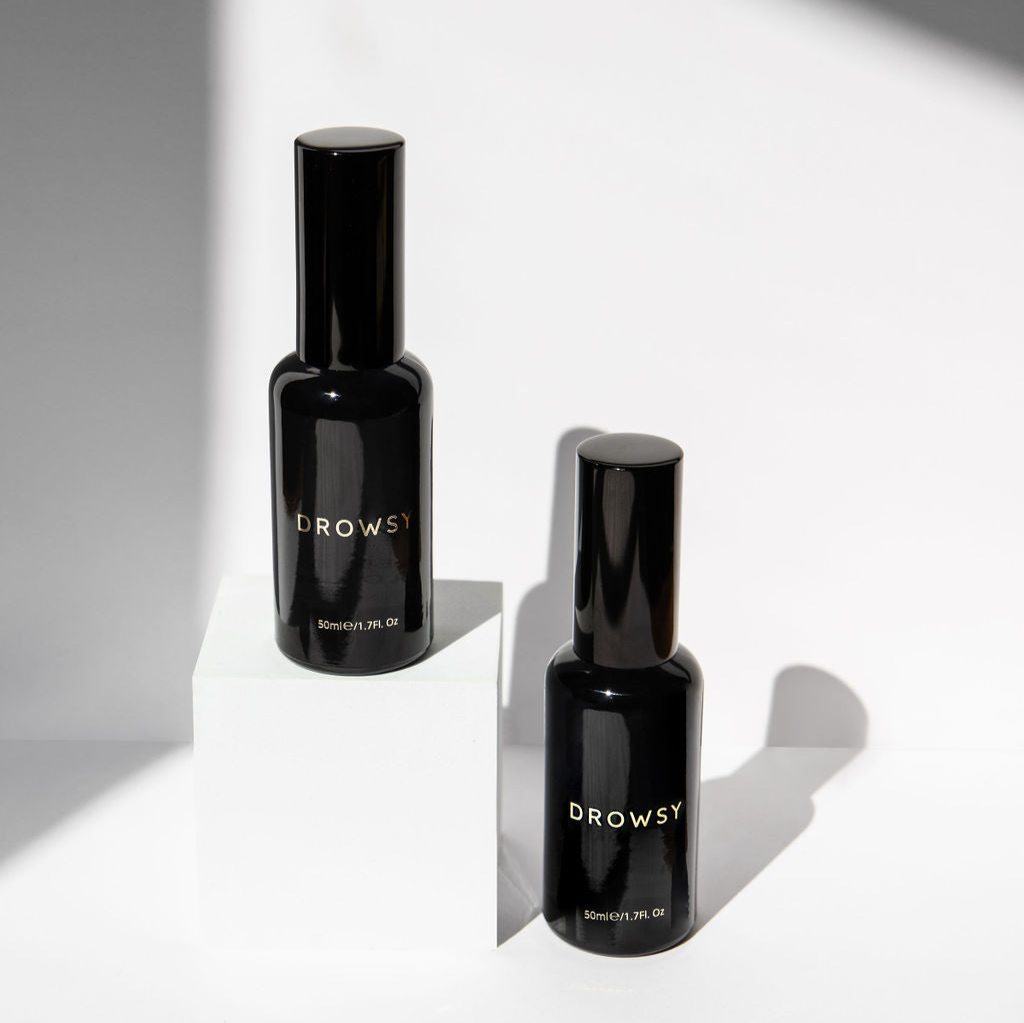 Two black glass bottles labeled Drowsy with gold text are displayed on a white surface; one bottle stands on a white block while the other is placed beside it, both casting soft shadows.