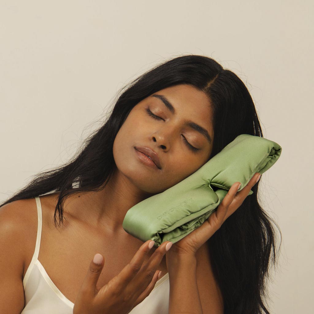 A woman with long dark hair and closed eyes rests her head gently on a light green satin pillow, appearing calm and relaxed against a plain background.