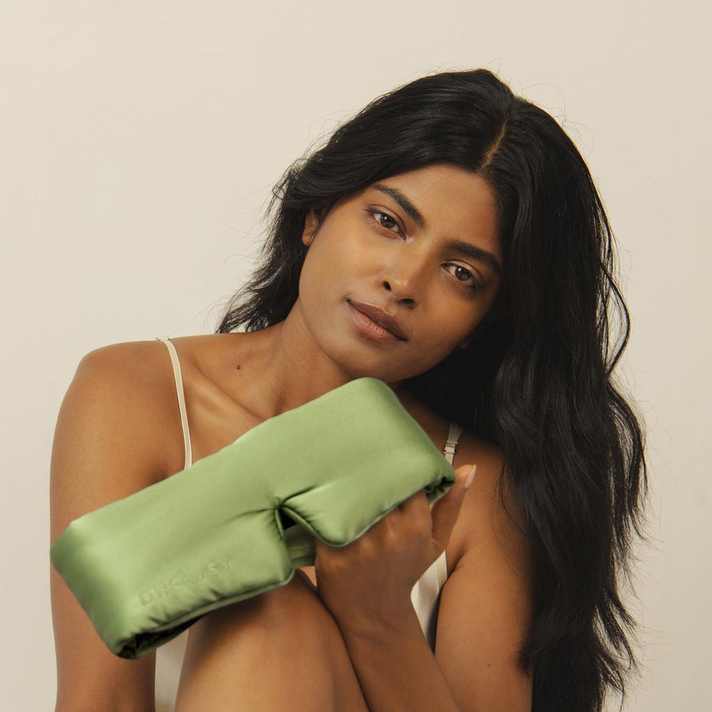 A woman with long dark hair in a white tank top gently holds the us.drowsysleepco Pistachio Green satin eye pillow, looking softly at the camera against a plain light background.