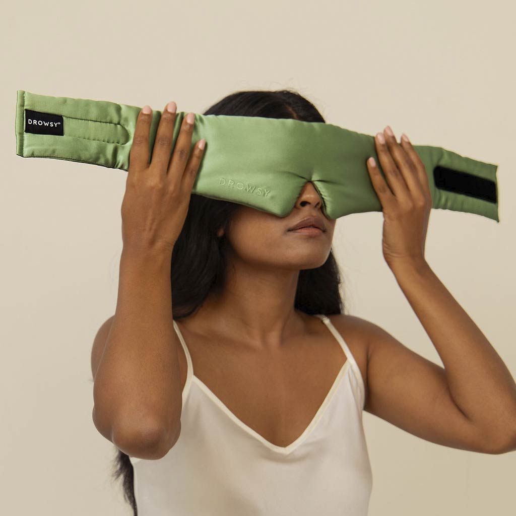 A woman in a white camisole holds the us.drowsysleepco Pistachio Green silk eye mask over her eyes against a plain beige background, embodying stylish beauty sleep.