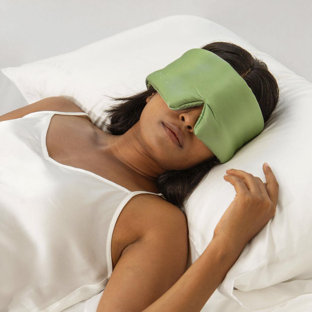 A woman in a white satin sleepwear lies on a bed with a white pillow, wearing a large green eye mask that covers her eyes and forehead. She appears to be relaxed or sleeping.