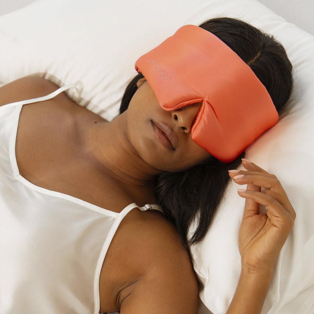 A woman in a white satin camisole lies on her back, wearing the us.drowsysleepco Summer Coral silk eye mask, which covers her eyes and nose for ultimate beauty sleep. Her right hand gently rests on the pillow beside her head.